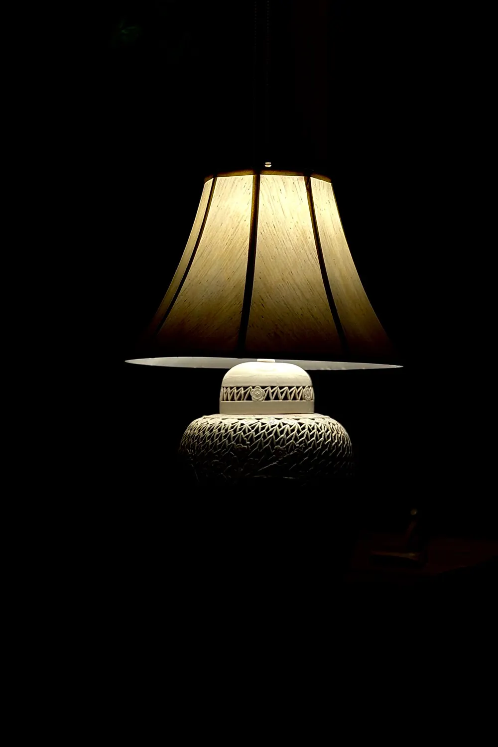 A table lamp with a pleated shade on a turned ceramic base, glowing alone in a dark room