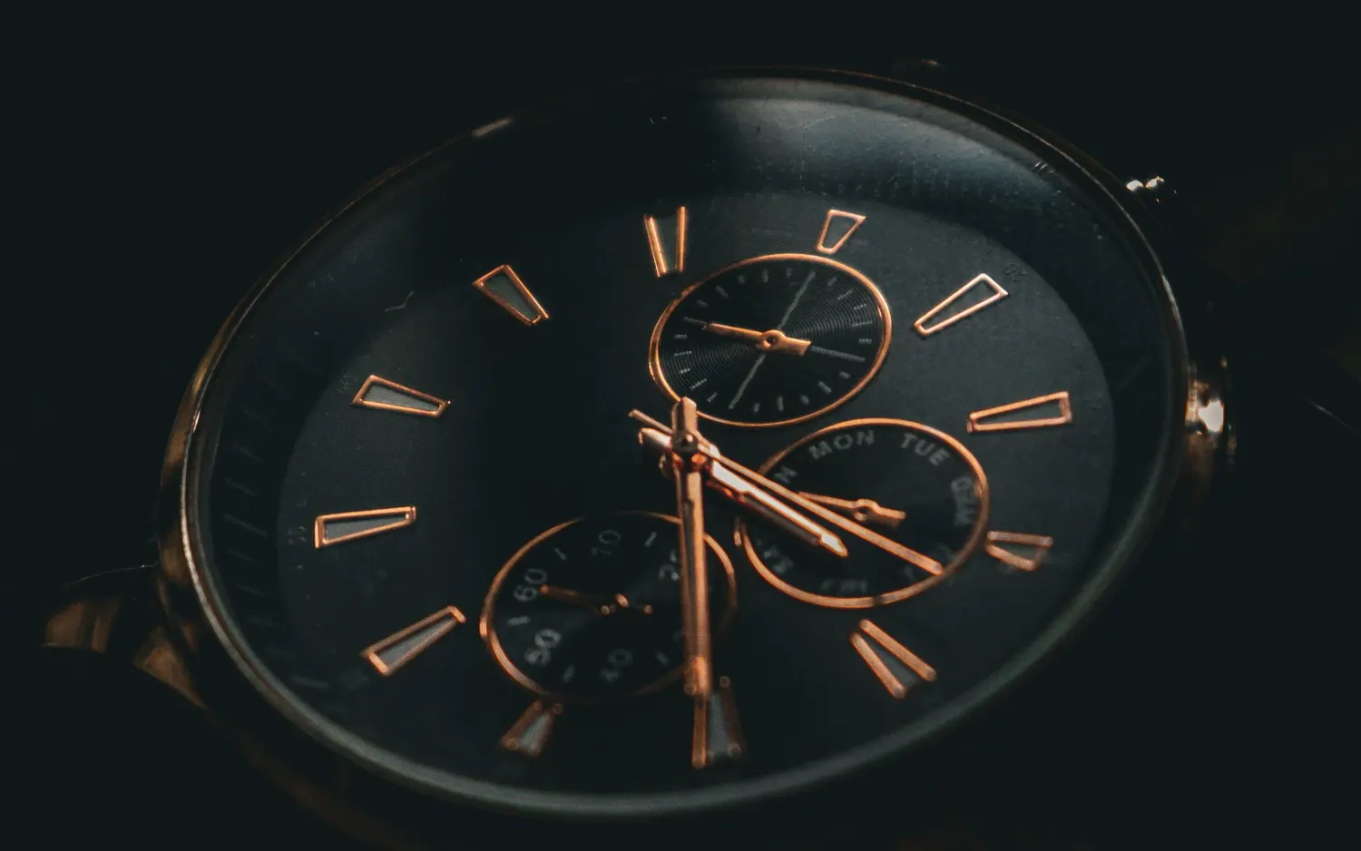 The black dial of a rose-gold chronograph wristwatch with three sub-dials, lit from one side in the dark