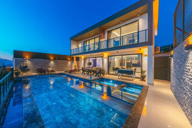 A modern two-storey villa at dusk, its glazed living room lit, with a lit pool in front.