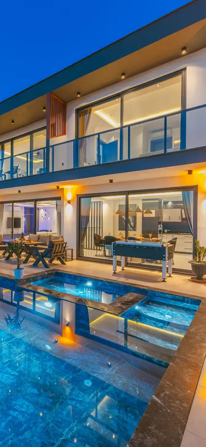 A modern villa at dusk with its living room lit and a lit pool in front.