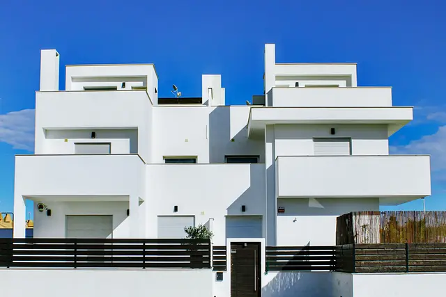 The white rendered facades of a terrace of new houses, with flat roofs and balconies, under a blue sky.