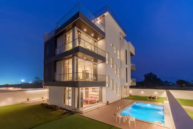 A modern white three-storey villa at twilight, every floor lit behind glass, with a lit pool in the garden.