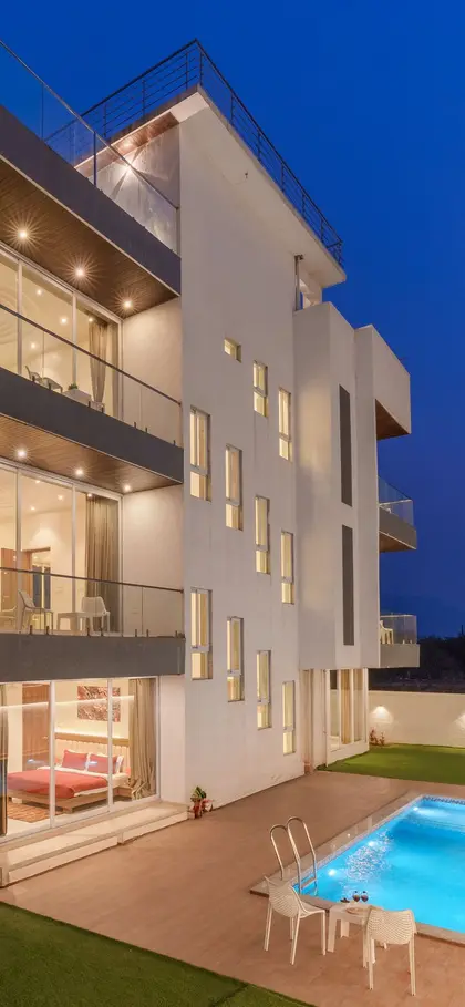 A modern white villa at twilight with its floors lit and a lit pool below.