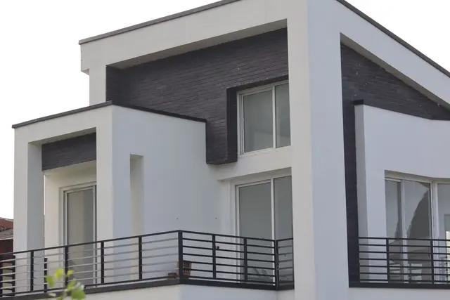 A modern white rendered house with a dark slate panel, large glazed doors and a black balcony rail, in daylight.