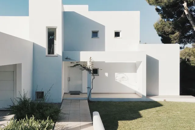 A white two-storey house with a flat roof and a pine tree, under a clear blue sky.